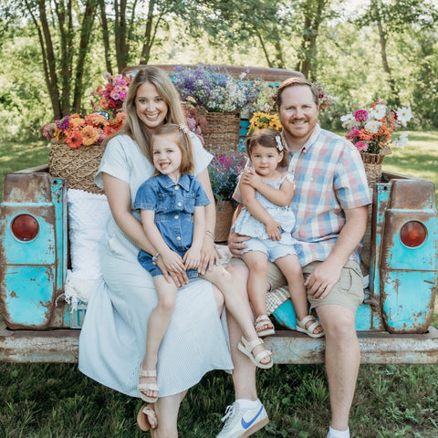 Family of four sitting in the back of a vintage truck with floral decorations in a natural setting.