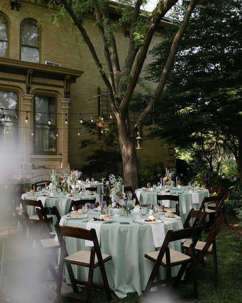 Outdoor wedding setup with tables and chairs under a large tree in front of a building.