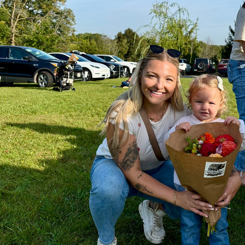 Woman and a child holding flowers at flower farm with cars and trees in the background