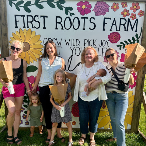 Group of people posing in front of 'First Roots Farm' sign with children holding wooden boxes.