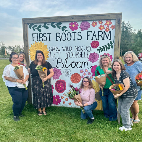Group of women holding flowers in front of a 'First Roots Farm' sign.