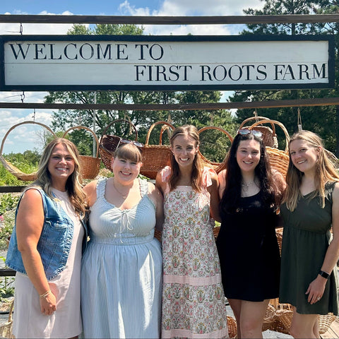 Five women posing in front of a 'Welcome to First Roots Farm' sign.
